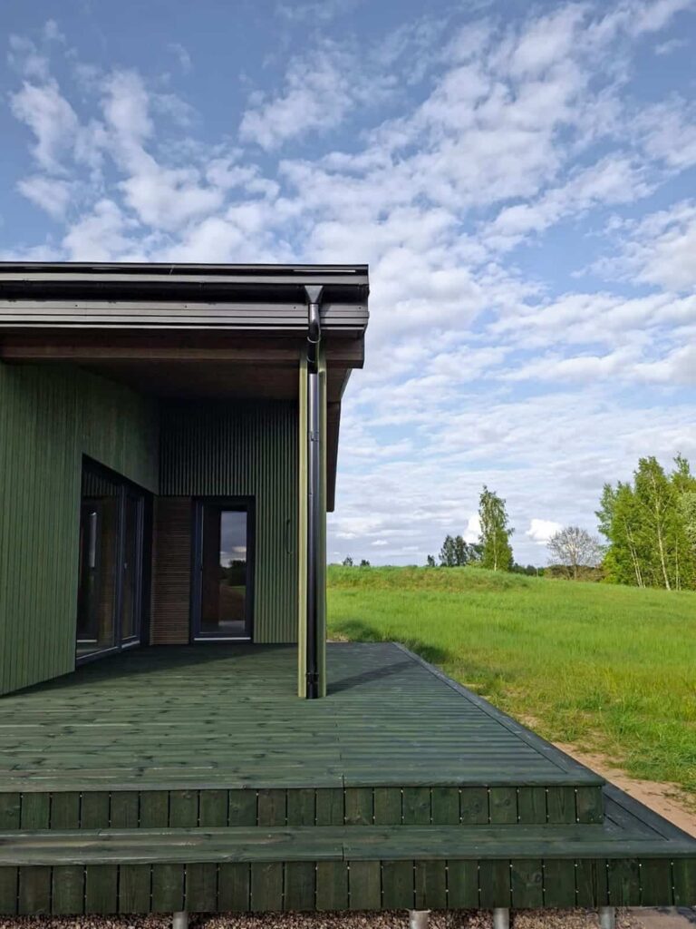 A corner of a terrace made of charred spruce, in Medeksa Moss color, with a green meadow and forest in the background.